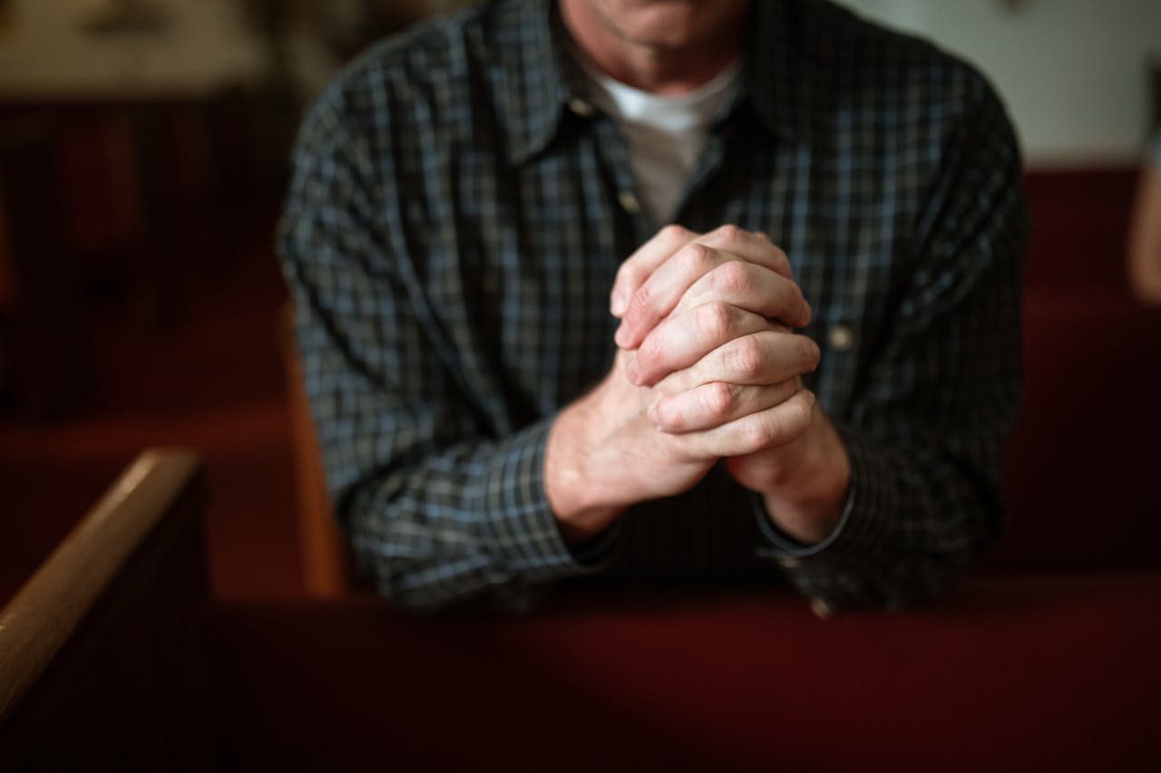brand-01 A man in a checked shirt praying in a church, hands clasped together.
