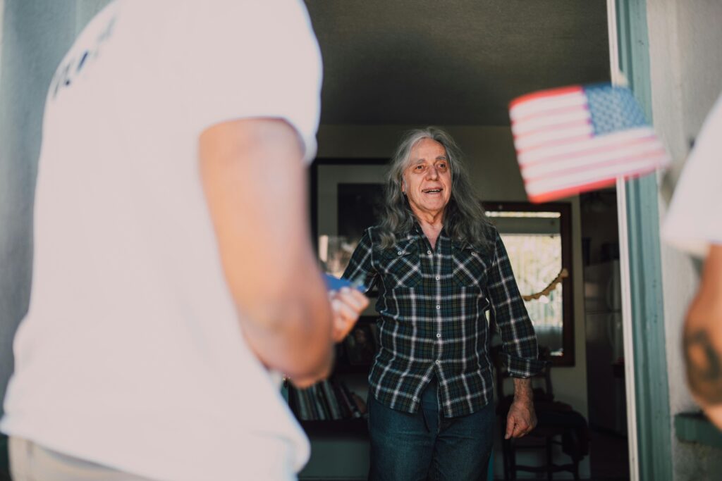 Elderly man welcomes volunteers at his front door, American flag in hand, symbolizing community service.